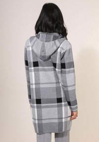 Cardigan à capuche à carreaux gris avec un mélange de noir, blanc et nuances de gris. Tissu doux, design mi-long et fentes sur les côtés pour faciliter les mouvements.