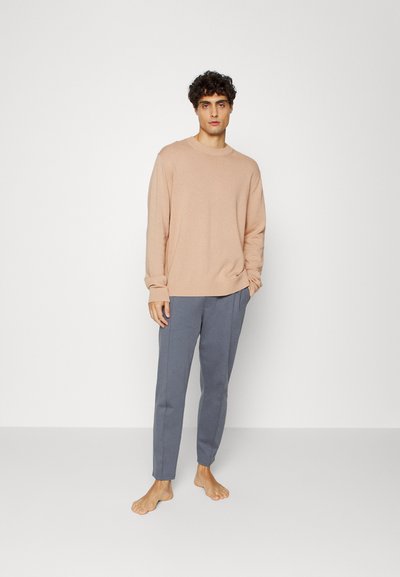 Suéter crew-neck de pêssego claro, com punhos canelados, combinado com calças cinzentas ajustadas, ambos em tecidos suaves. O modelo está descalço contra um fundo simples.