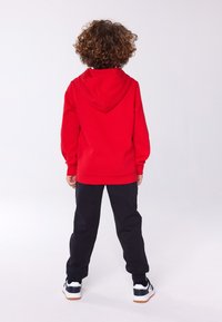 Red hooded sweatshirt with a relaxed fit, black sweatpants, and white athletic shoes with navy accents. Back view shows curly hair and simple design.