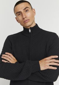 Sort strikket zip-up sweater med høj krave, der har strukturerede mønstre på skuldre og ærmer. Glatte manchetter og lynlåsdetalje.