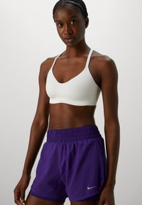 En atletisk kvinne iført en hvit sports-BH og lilla Nike løpeshorts, stående mot en enkel lys bakgrunn.