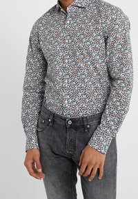 Camicia bianca a maniche lunghe con un motivo floreale multicolore, con accenti blu, rossi e verdi, abbinata a jeans grigi.