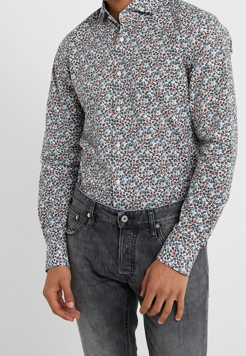 Camicia bianca a maniche lunghe con un motivo floreale multicolore, con accenti blu, rossi e verdi, abbinata a jeans grigi.