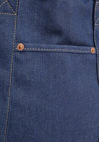 Tmavě modrá denímová tkanina s diagonálním žebrováním, obsahující měděné akcenty a viditelné zlaté švy. Zahrnuje detail náprsní kapsy.