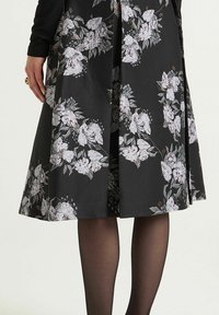 Jupe noire évasée avec imprimé floral en rose clair et blanc, en tissu texturé avec un design plissé. Collants noirs visibles.