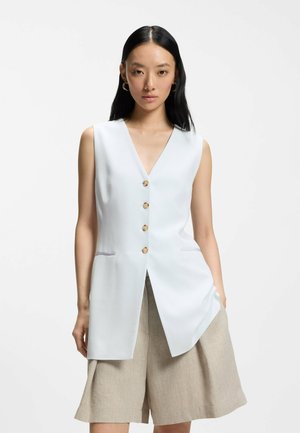 Donna con lunghi capelli neri indossa un gilet bianco senza maniche con bottoni e pantaloncini beige a gamba larga, in piedi davanti a uno sfondo chiaro uniforme.