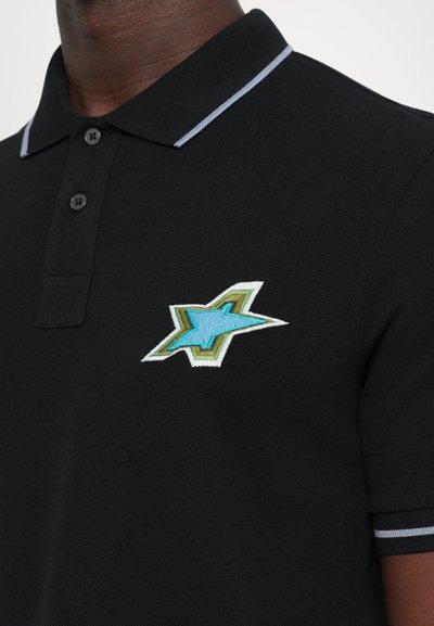 Crna polo majica s svijetloplavim i zelenim izvezenim logotipom u obliku zvijezde na prsima, nosi ju osoba tamne puti.