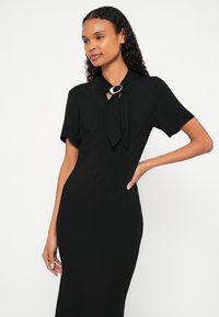 Femeie într-o rochie neagră mulată cu mâneci scurte și o broșă decorativă la decolteu, stând pe un fundal alb.