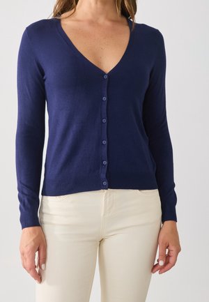 Femme portant un cardigan bleu marine ajusté à col en V boutonné et un pantalon couleur crème, debout devant un fond gris clair.