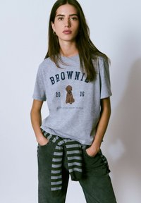 Camiseta de algodón gris con un gráfico de un perro marrón y el texto "BROWNIE 2006". Combinada con pantalones oscuros y un suéter a rayas atado a la cintura.