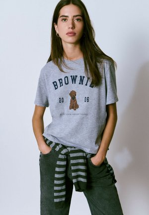 Camiseta de algodón gris con un gráfico de un perro marrón y el texto "BROWNIE 2006". Combinada con pantalones oscuros y un suéter a rayas atado a la cintura.