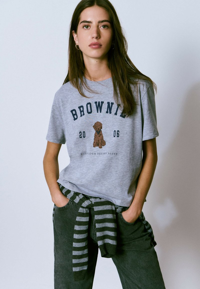 Camiseta de algodón gris con un gráfico de un perro marrón y el texto "BROWNIE 2006". Combinada con pantalones oscuros y un suéter a rayas atado a la cintura.