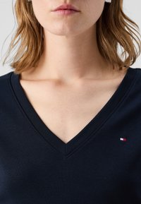 Námořnický svetr s véčkovým výstřihem z měkké látky má malé logo s červenými, bílými a modrými akcenty na levé straně blízko lemu.
