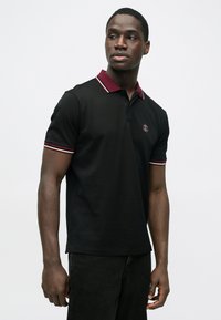 Svart poloshirt med en burgunderkrage og stripete detaljer. Har en logo på venstre bryst, laget av et glatt, lett stoff.