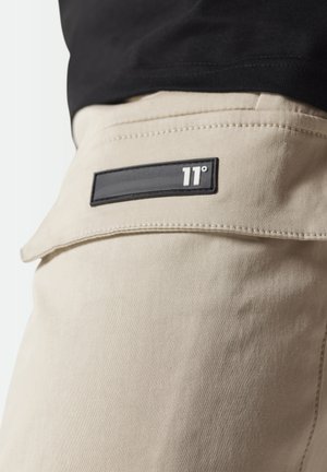 Pantalones beige con una textura suave, detalles cosidos y una etiqueta rectangular negra con letras blancas que dice "11°" en el bolsillo trasero.