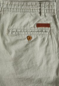 Shorts en tissu gris clair avec une poche arrière à fermeture boutonnée, des détails cousus et un écusson de marque en cuir marron.
