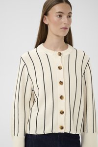 Tânără îmbrăcată într-un cardigan crem cu dungi negre verticale și nasturi mari maro, purtând pantaloni închiși la culoare, pe un fundal simplu.