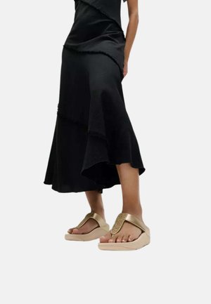 Persona que lleva una falda negra asimétrica y sandalias de plataforma beige con tiras anchas y detalle brillante sobre un fondo blanco.