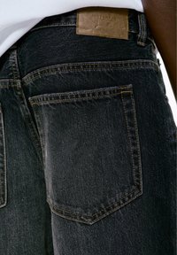 Prim-plan al buzunar din spate al unei perechi de blugi negri din denim, cu cusături maro și etichetă din piele a brandului, purtați cu o cămașă albă.
