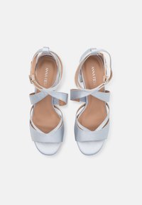 Sandales en satin bleu clair avec plusieurs brides croisées, bout ouvert et un talon bloc de hauteur moyenne. La semelle intérieure affiche le logo "ANNA FIELD".