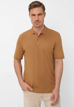 Man met een bruin poloshirt met korte mouwen en knoopsluiting, en lichtbeige broek, staand met één hand in de zak.