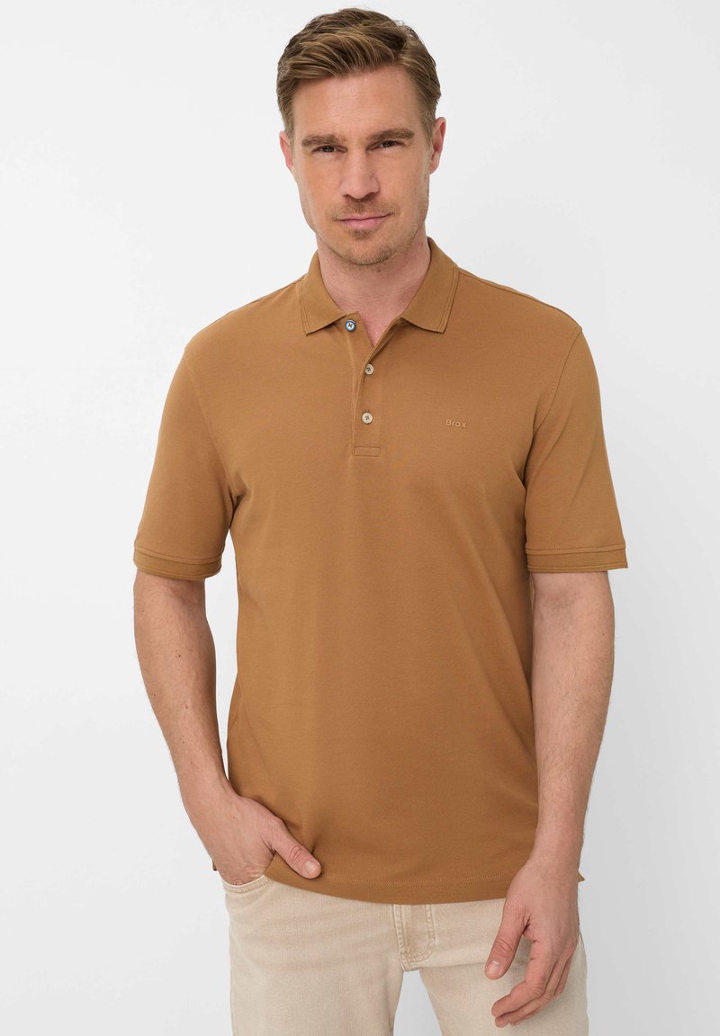 Mann trägt ein braunes Poloshirt mit kurzen Ärmeln und Knopfleiste sowie hellbeige Hosen, steht mit einer Hand in der Tasche.