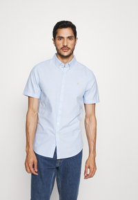 Camicia azzurro chiaro a maniche corte, con colletto a punta, un piccolo logo sul petto e una vestibilità ampia. Indossati sotto jeans di denim.