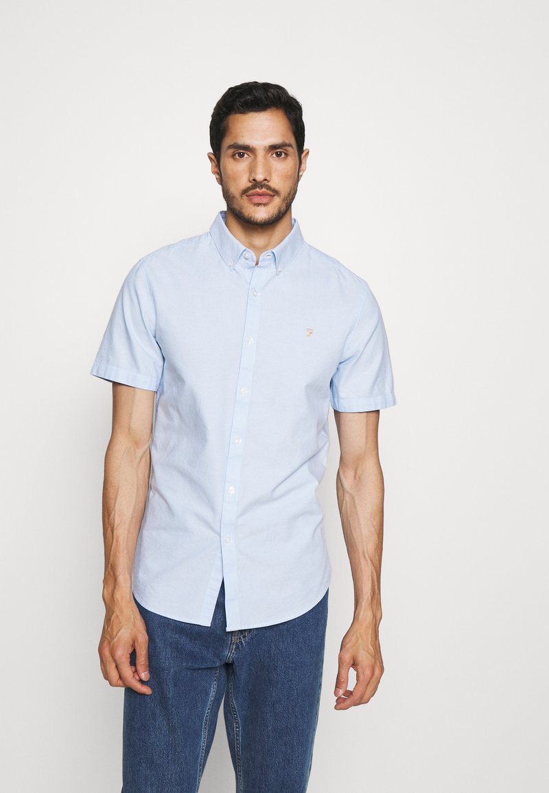 Camicia azzurro chiaro a maniche corte, con colletto a punta, un piccolo logo sul petto e una vestibilità ampia. Indossati sotto jeans di denim.