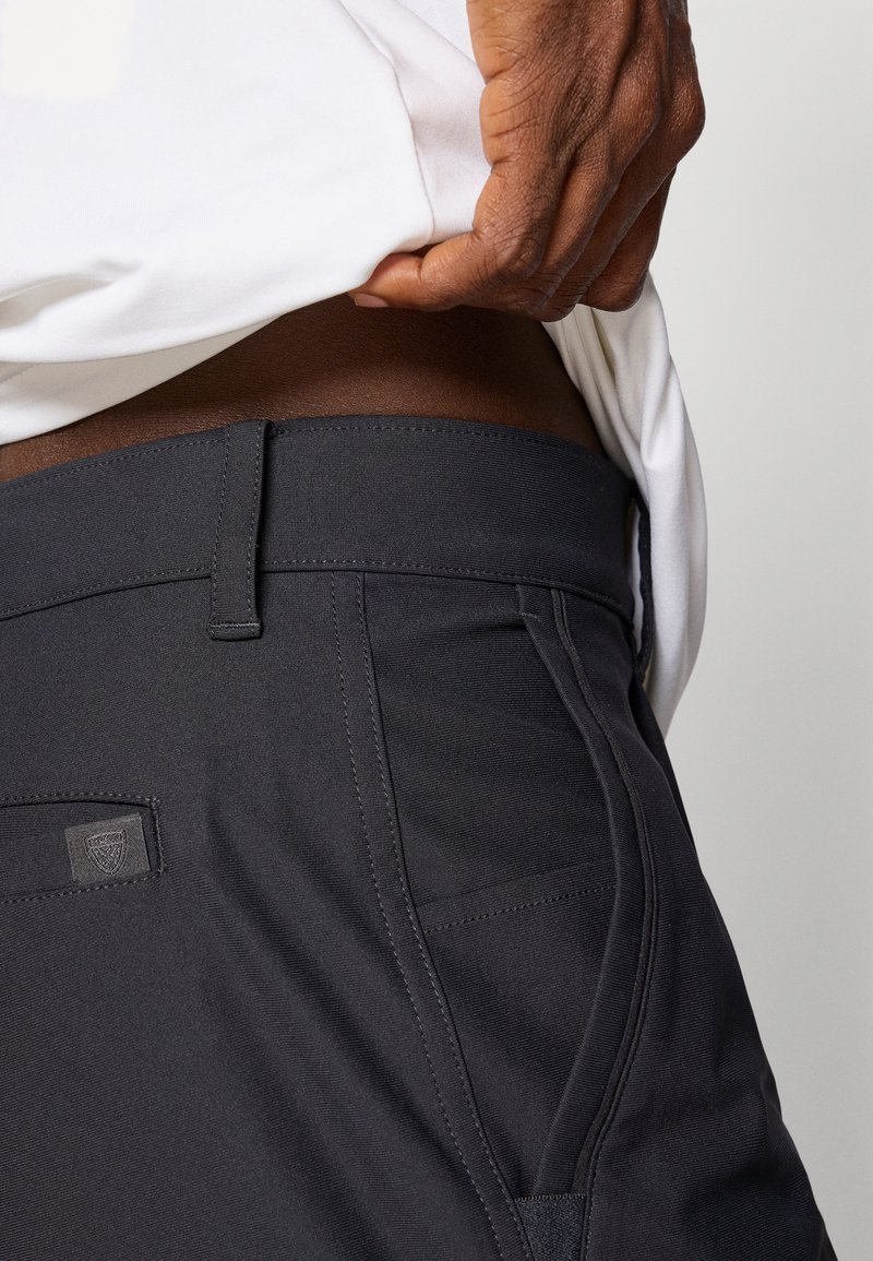 Pantaloni grigi scuri con una texture liscia, dotati di una vita aderente, tasche laterali e un piccolo logo sul retro. Design semplice.