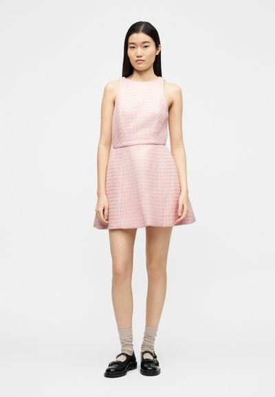 Νεαρή γυναίκα που στέκεται με ένα ροζ αμάνικο φόρεμα με υφή, συνδυασμένο με μπεζ κάλτσες και μαύρα παπούτσια Mary Jane σε λευκό φόντο.