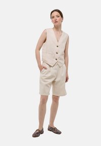 Gilet en lin avec un décolleté en V profond et trois boutons, associé à un short longueur genou de la même teinte claire, stylisé avec des chaussures à motifs à enfiler.