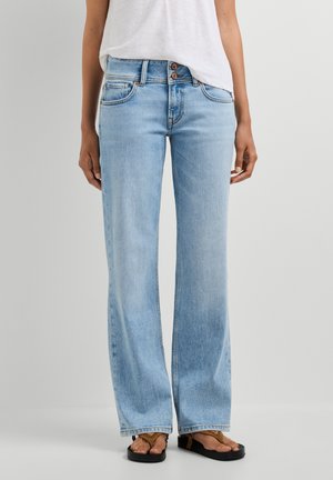 Person iført lyseblå jeans med vide ben, hvid top og brune sandaler, stående mod en ensfarvet lys baggrund.