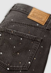 Donkerdenim jeans met een gestructureerd oppervlak, versierd met verspreide gouden en zilveren accenten, en een tan leren merklabel op de tailleband.