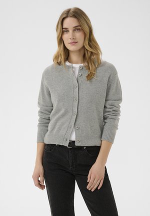 Ung kvinde med langt blondt hår iført en grå knappet cardigan over en hvid skjorte og sorte jeans står mod en hvid baggrund.
