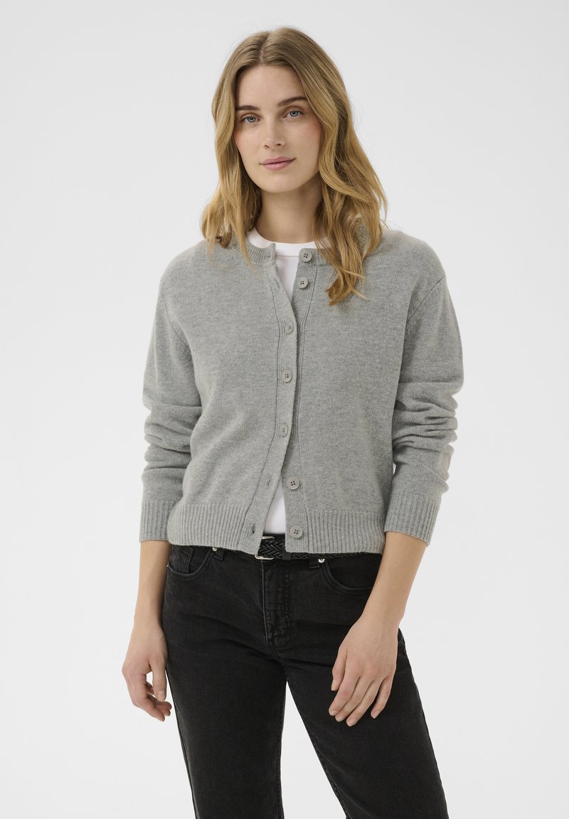 Jeune femme aux longs cheveux blonds portant un cardigan gris boutonné par-dessus une chemise blanche et un jean noir, debout contre un fond blanc.