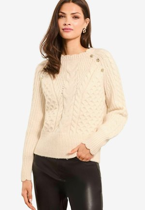 Maglione a maglia di colore crema con motivi a treccia, polsini e orlo ribattuti, ed eleganti bottoni decorativi sulle spalle. Ancora in primo piano.