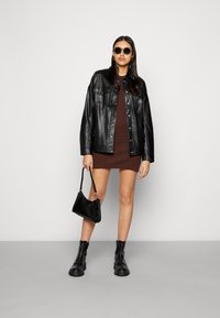 Jaqueta de couro preta com botões de pressão sobre um vestido justo castanho. Os acessórios incluem óculos de sol pretos e uma pequena bolsa texturizada. Botas pretas chunky.