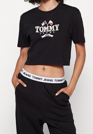 Maglietta nera corta con logo ricamato "TOMMY JEANS" accompagnato da bandiere, abbinata a pantaloni neri e una cintura elastica bianca.