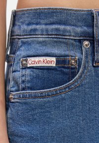 Jeans de ganga azul escuro com uma vista aproximada do bolso da frente. Apresenta uma etiqueta "Calvin Klein" em vermelho e branco e detalhes em prata.