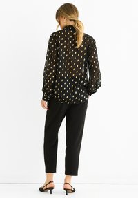 Black sheer blouse with gold teardrop patterns, long sleeves, and a relaxed fit. Paired with tapered black trousers and open-heel sandals.