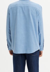 Chemise bleu clair à manches longues avec col pointu, texture lisse et poignets boutonnés. L'ourlet est légèrement arrondi en bas. Jean bleu visible.