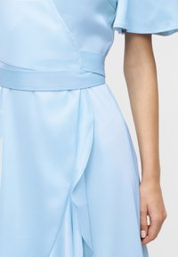 Φόρεμα ανοιχτού γαλάζιου χρώματος με εφαρμοστή μέση, επικαλυπτόμενο μπροστινό πάνελ και κοντά μανίκια με βολάν, που απεικονίζεται στον κορμό και το χέρι ενός ατόμου πάνω σε ουδέτερο φόντο.