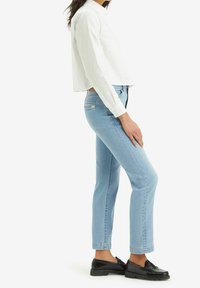 Žena v světle modrých slim džínách, černých mokasínech a bílém cropped tričku s dlouhým rukávem, stojící v profilu na bílém pozadí.