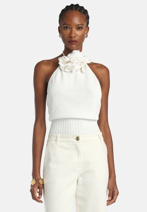 Fonott hajú nő fehér ujjatlan kötött felsőt visel fodros gallérral, krémszínű magas derekú nadrágot, arany fülbevalót és karkötőt, előre néz.