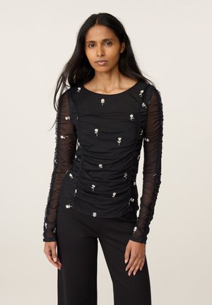 Vrouw met lang donker haar die een zwarte doorschijnende lange mouwen top met witte rozenpatronen en een zwarte broek draagt, staand voor een effen achtergrond.