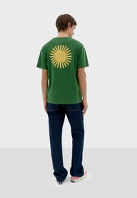 Camiseta verde de manga corta con un gran gráfico de un sol amarillo en la espalda. Combinada con pantalones de mezclilla azules y zapatillas multicolores.
