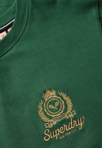Sweatshirt en coton vert avec un col rond et un logo brodé doré proéminent représentant une couronne et des raquettes croisées.