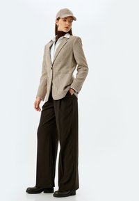 Een geblokte blazer in beige en bruin, gecombineerd met een bruine coltrui en brede zwarte broek. Accessoires omvatten een bijpassende geblokte pet.