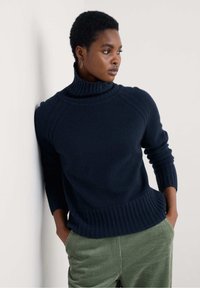 Mornarsko turtleneck pulover z rebrastim ovratnikom in obročki, mehak teksturiran, prilegajoč silhuet, v kombinaciji z zelenimi kordnimi hlačami.