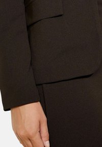 Blazer nero in tessuto con una texture liscia. Presenta un collo strutturato e una tasca frontale. La mano è leggermente appoggiata sul manico.
